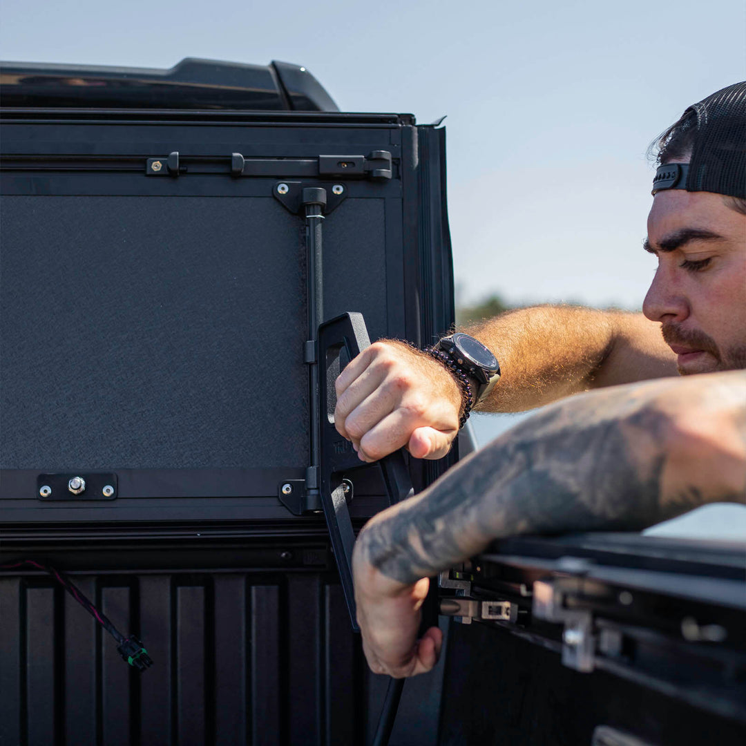 Person operating the TruxArmor QuadAxis Pro flip-up tonneau cover support handle on a Toyota Tacoma truck bed.